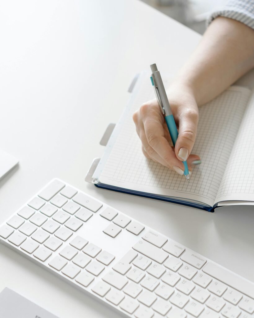 pexels-photo-8572606-8572606 Close-up of a hand writing in a notebook next to a white keyboard on a desk.
